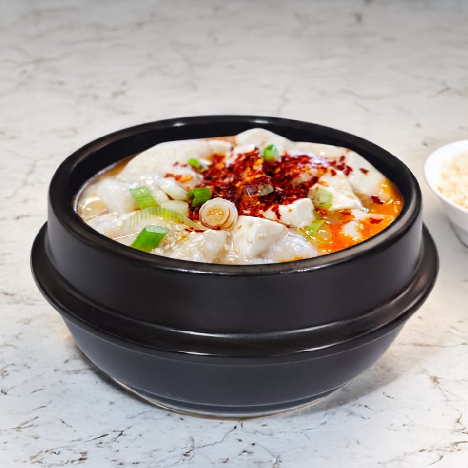 A black bowl of spicy tofu and fish stew, garnished with chili flakes and green onions.